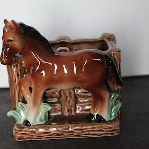 May include: A brown ceramic horse planter with a dark brown mane and tail. The horse is positioned in front of a wooden fence-like structure. Green and white accents are at the base. A decorative item for home decor.