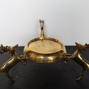 May include: A brass candle holder with a circular base supported by two deer figures. A third deer figure rises from the center, holding a small platform. The candle holder is a decorative item.