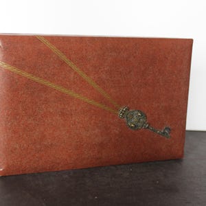 May include: A rectangular, reddish-brown box with a textured surface. The box is decorated with a gold-colored key design. The key is ornate and detailed, with a vintage aesthetic. The box is likely used for storage or display.