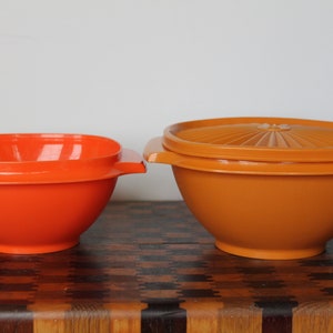 May include: Two plastic mixing bowls, one orange and one brown. The orange bowl has a square shape and the brown bowl has a round shape.