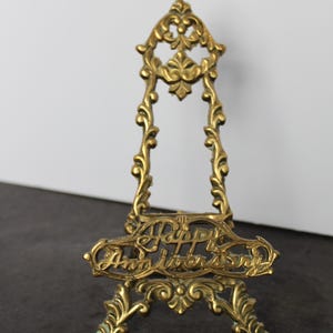 May include: A gold-toned metal easel stand featuring an ornate, scrollwork design. The front horizontal bar displays the embossed text "Happy Anniversary" in a cursive script. The structure has a pointed top and a wide, decorative base.