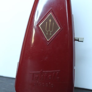 May include: A maroon Taktell piccolo metronome. The metronome has a diamond-shaped emblem with a gold-colored design. The word "Taktell" and "piccolo" are printed on the front. It has a winding key and a small foot.
