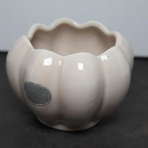 May include: A small, pale pink ceramic bowl with a scalloped, flower-like rim and a rounded, ribbed body. A silver-colored oval sticker is affixed to the side of the exterior, featuring faint, illegible embossed text on its surface.