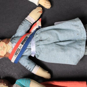 May include: A handmade doll wearing a blue dress with a white sash and a red, white, and blue sash that says "Votes for Women".