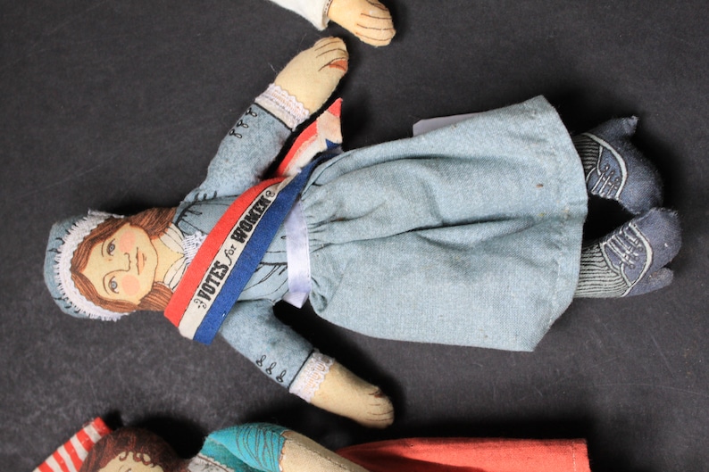May include: A handmade doll wearing a blue dress with a white sash and a red, white, and blue sash that says "Votes for Women".