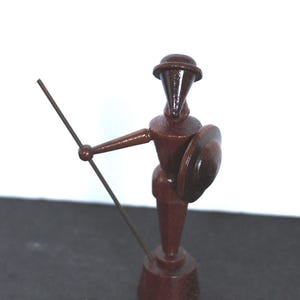 May include: A small, wooden figurine of a matador in a dark brown finish. The figure holds a spear in one hand and a shield in the other. The matador wears a hat and stands on a small base. The figurine is a decorative item.