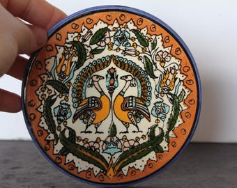 May include: A round, hand-painted ceramic plate with a vibrant design. The plate features two stylized birds facing each other, surrounded by floral and leaf motifs in orange, yellow, blue, and green. The rim is a deep blue.