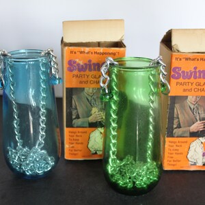 May include: Two vintage Swinger party glasses with chains, one teal and one green, with original packaging. The boxes are orange with the text "Swinger Party Glass and Chain". The glasses are designed to hang around the neck.
