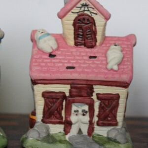 May include: A pink and white ceramic house with a red roof and brown doors. There are three white ceramic figures on the roof and one in the doorway.