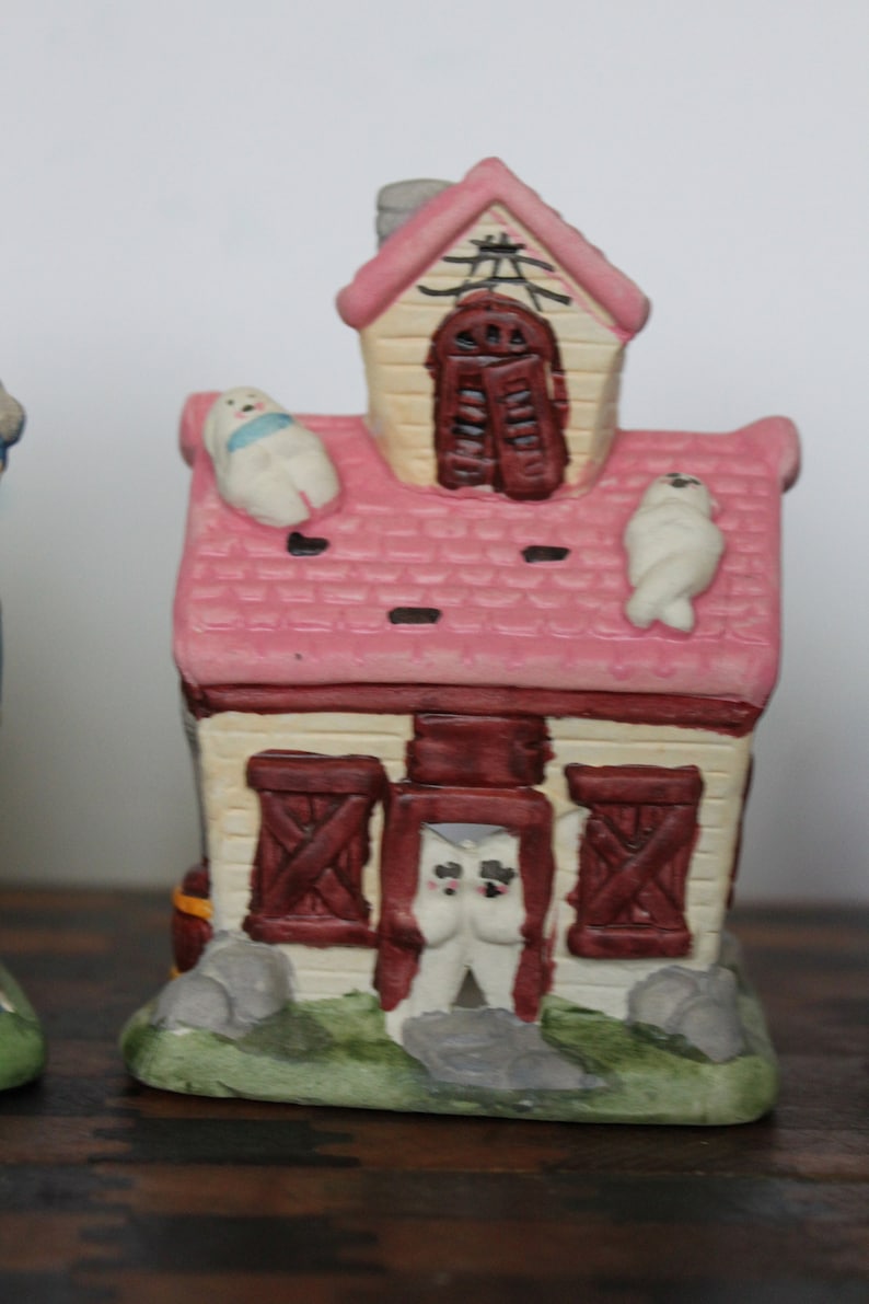 May include: A pink and white ceramic house with a red roof and brown doors. There are three white ceramic figures on the roof and one in the doorway.