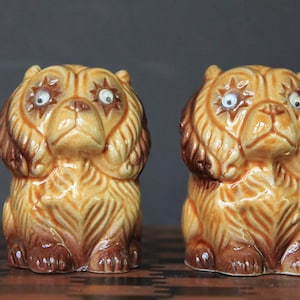 May include: A pair of ceramic salt and pepper shakers shaped like brown and tan dogs with white eyes. The dogs have a detailed design and are sitting upright.