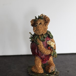 May include: A brown teddy bear figurine wearing a red strawberry costume with green leaves and holding a bouquet of white flowers. The bear is standing on a green leaf base.