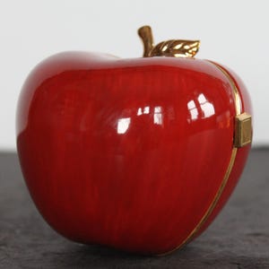 May include: A glossy red apple-shaped decorative box with a gold-colored stem and leaf. The box has a gold-colored trim and clasp. The apple is a vibrant red color and reflects light.