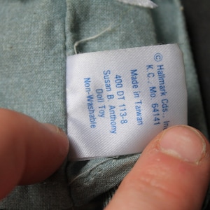 May include: A white tag with blue text attached to a light blue fabric. The tag reads "Hallmark Cds., Inc. K.C., Mo. 64141 Made in Taiwan 400 DT 113-8 Susan B. Anthony Doll Toy Non-Washable"