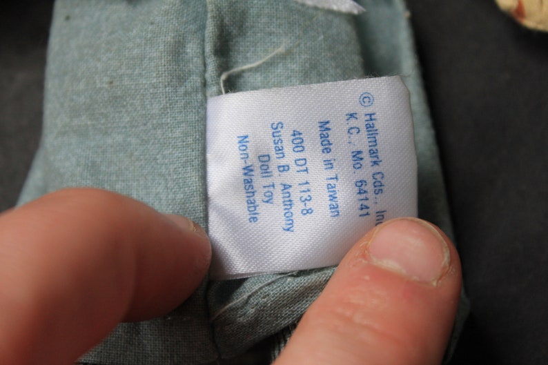 May include: A white tag with blue text attached to a light blue fabric. The tag reads "Hallmark Cds., Inc. K.C., Mo. 64141 Made in Taiwan 400 DT 113-8 Susan B. Anthony Doll Toy Non-Washable"