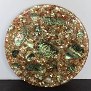 May include: A circular coaster featuring a clear resin base embedded with a dense mixture of small, crushed orange and tan stone fragments, interspersed with several larger, iridescent abalone shell pieces that display green and blue hues.