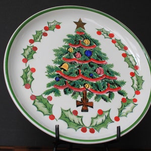 May include: A white ceramic plate with a green rim features a hand-painted Christmas tree design. The tree is decorated with red ribbons, green leaves, and colorful ornaments. The plate is surrounded by a wreath of holly leaves and red berries.