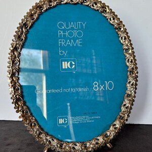 May include: An oval gold-toned photo frame with a teal blue background. The frame has an ornate, floral-like design around the edge. Text on the frame reads "QUALITY PHOTO FRAME by IIC" and "Guaranteed not to tarnish 8x10".