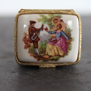 May include: A small, rectangular trinket box with a gold-colored metal frame. The lid features a painted scene of a man playing a guitar for a woman. The woman is seated, and the scene is set in a garden. The box is likely vintage.