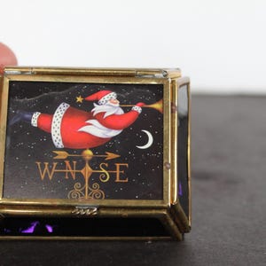 May include: A small, square, vintage trinket box with a gold-colored metal frame. The box features a Christmas-themed image of Santa Claus flying across a starry night sky, with a crescent moon and a compass rose design.