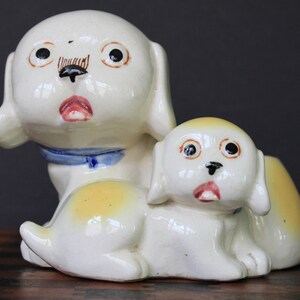 May include: A white ceramic figurine of two dogs. The larger dog has a blue collar and yellow accents. The smaller dog is sitting on the larger dog's back. Both dogs have black eyes and painted details.
