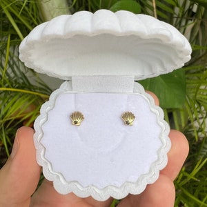 May include: A pair of small gold-toned stud earrings shaped like seashells, displayed inside a white, velvet-textured jewelry box that is molded into the shape of an open clam shell with a scalloped edge and a white interior insert.