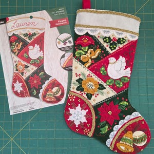 May include: A felt Christmas stocking with a patchwork design, featuring a dove, poinsettias, and bell ornaments. The stocking is 18 inches (45.72 cm) long and has a white cuff with gold trim and a red hanging loop. The kit includes beads.