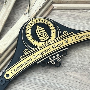 May include: A black and gold wooden hanger with the words "United States Army" and "Command Sergeant Major W.J. Chavez". The hanger features a gold emblem and stars.