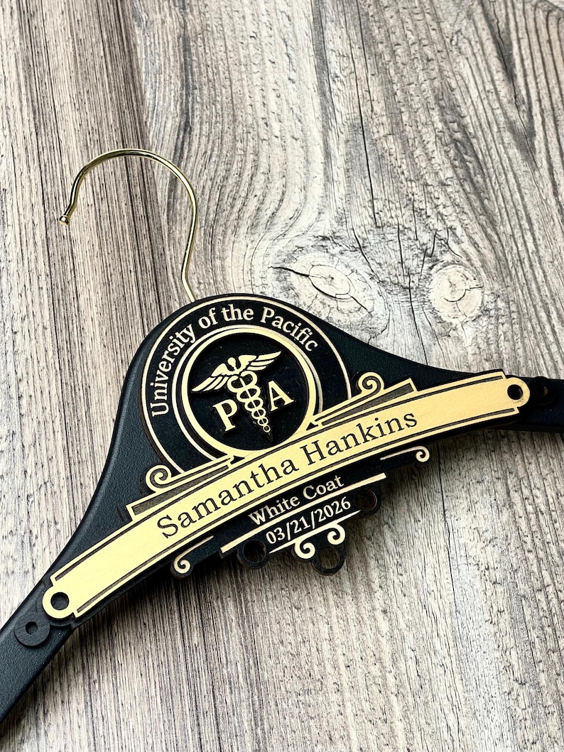 May include: A black clothes hanger with a gold metal hook. The hanger features a circular emblem with the text University of the Pacific and a caduceus symbol. A gold plaque displays the name Samantha Hankins, White Coat, and 03/21/2026.