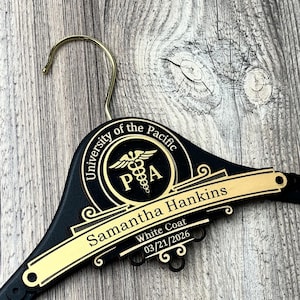 May include: A black clothes hanger with a gold-colored metal hook. The hanger features a circular emblem with the text University of the Pacific and a caduceus symbol with the letters PA. A gold plaque displays the name Samantha Hankins, White Coat, 03/21/2026.