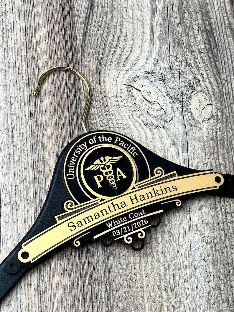 May include: A black clothes hanger with a gold-colored metal hook. The hanger features a circular emblem with the text University of the Pacific and a caduceus symbol with the letters PA. A gold plaque displays the name Samantha Hankins, White Coat, 03/21/2026.