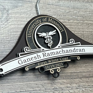 May include: A wooden coat hanger with a black and gold design. The hanger has a circular emblem with the text "University of Missouri" and a medical symbol. Below the emblem is a banner with the text "Ganesh Ramachandran First White Coat 10/19/2023".