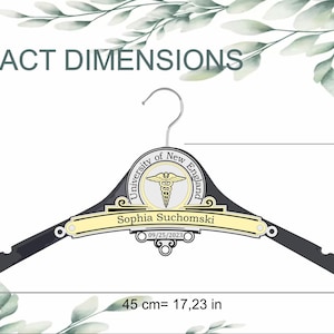 May include: A black clothes hanger featuring a central emblem with the text University of New England and a caduceus symbol. A yellow nameplate reads Sophia Suchomski with the date 09/25/2023. Dimensions are 17.23 inches wide, 6.19 inches high.