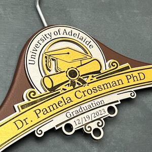 May include: A brown wooden hanger with a gold and silver design. The design features a graduation cap, a scroll, and the text "University of Adelaide, Dr. Pamela Crossman PhD, Graduation 12/19/2023".