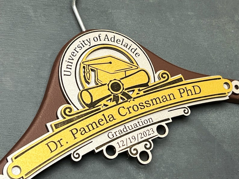 May include: A brown wooden hanger with a gold and silver design. The design features a graduation cap, a scroll, and the text "University of Adelaide, Dr. Pamela Crossman PhD, Graduation 12/19/2023".