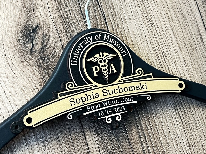 May include: A black wooden clothes hanger featuring a gold-colored plaque with the text University of Missouri, a caduceus symbol, the letters PA, the name Sophia Suchomski, the phrase First White Coat, and the date 10/19/2023 engraved in black.