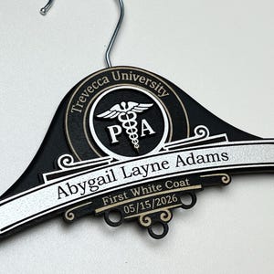 May include: A black wooden clothes hanger featuring a circular emblem with the text Trevecca University and a caduceus symbol with the letters PA. A silver plaque displays the name Abygail Layne Adams, First White Coat, and 05/15/2026.