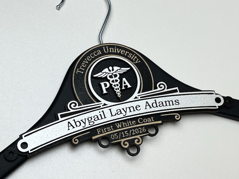 May include: A black wooden clothes hanger featuring a circular emblem with the text Trevecca University and a caduceus symbol with the letters PA. A silver plaque displays the name Abygail Layne Adams, First White Coat, and 05/15/2026.