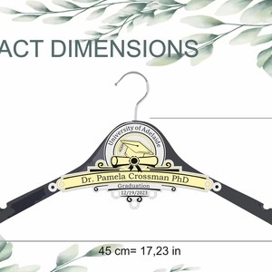 May include: A black wooden hanger with a yellow and gray University of Adelaide graduation design. The hanger is 17.23 inches long, 6.19 inches high, and 3.3 inches wide. The design includes a graduation cap, diploma, and the text "Dr. Pamela Crossman PhD" and "Graduation 12/19/2023".