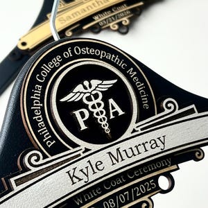 May include: Two black wooden clothes hangers featuring gold and silver metallic accents. Each hanger displays the Philadelphia College of Osteopathic Medicine logo, a caduceus symbol, and the names Kyle Murray and Samantha Hankins with dates.