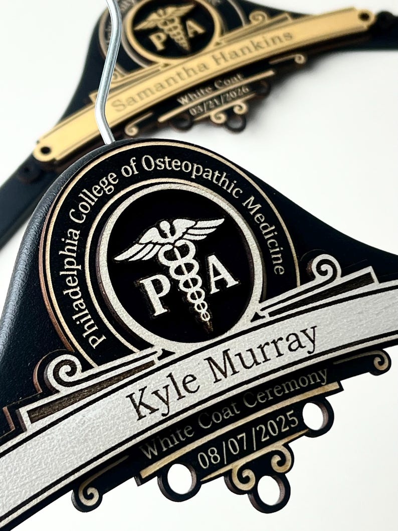 May include: Two black wooden clothes hangers featuring gold and silver metallic accents. Each hanger displays the Philadelphia College of Osteopathic Medicine logo, a caduceus symbol, and the names Kyle Murray and Samantha Hankins with dates.
