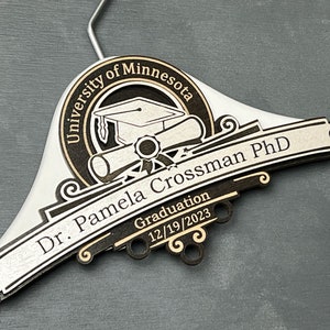 May include: Personalized PhD Hanger, PhD Gift, Personalized PhD Graduation Gift, Doctorate Graduation Gift, Hooding Ceremony Gift, Doctoral Candidate A white wooden hanger with a black and gold engraved design. The design features a graduation cap, diploma, and the text "University of Minnesota, Dr. Pamela Crossman PhD, Graduation 12/19/2023".