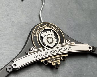 May include: A black wooden clothes hanger featuring a circular emblem with the text Cleveland Police and a central shield icon. A silver-toned horizontal plaque is attached, engraved with the name Officer Englehardt and the text Law & Order.