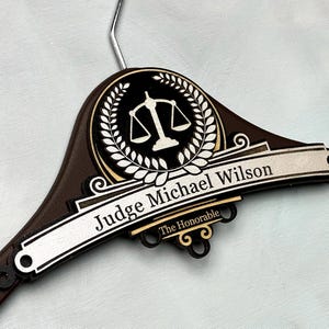 Può includere: Una gruccia in legno marrone scuro con un emblema circolare raffigurante un'icona bianca della bilancia della giustizia circondata da una corona d'alloro. Una targa orizzontale riporta il testo Judge Michael Wilson e The Honorable in bianco.
