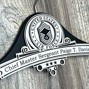 May include: Black wooden hanger with a silver emblem featuring the United States Air Force logo and the text "United States Air Force". Below the emblem is a silver banner with the text "Chief Master Sergeant Paige T. Davis" and three stars.
