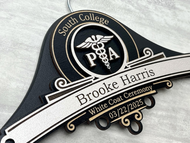 May include: A black wooden clothes hanger featuring a circular emblem with a caduceus symbol and the text South College PA. The hanger is engraved with the name Brooke Harris, the text White Coat Ceremony, and the date 03/22/2025 in white lettering.