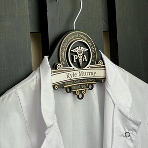 May include: A black wooden clothes hanger featuring a custom engraved plaque with the Philadelphia College of Osteopathic Medicine logo, the name Kyle Murray, the text White Coat Ceremony, and the date 08/07/2025, holding a white lab coat.