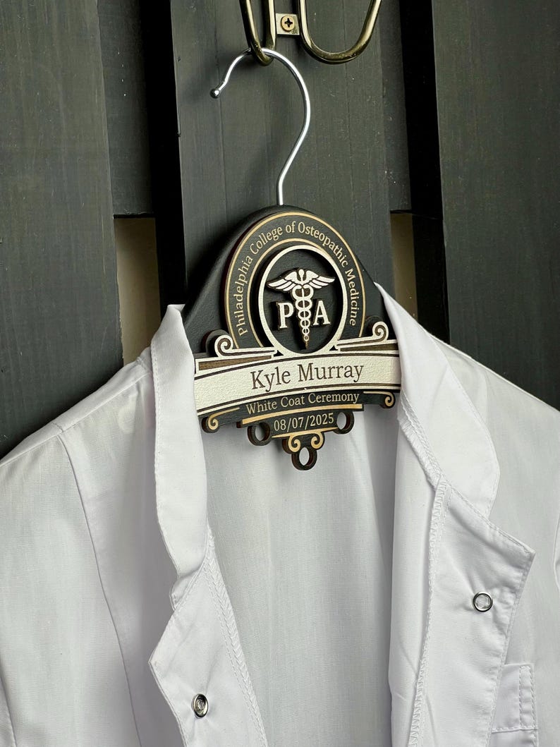 May include: A black wooden clothes hanger featuring a custom engraved plaque with the Philadelphia College of Osteopathic Medicine logo, the name Kyle Murray, the text White Coat Ceremony, and the date 08/07/2025, holding a white lab coat.