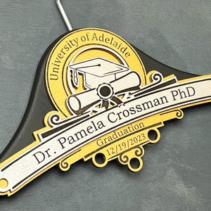 May include: Personalized PhD Hanger, PhD Gift, Personalized PhD Graduation Gift, Doctorate Graduation Gift, Hooding Ceremony Gift, Doctoral Candidate Black wooden hanger with a gold and silver inscription. The inscription reads "University of Adelaide, Dr. Pamela Crossman PhD, Graduation 12/19/2023". The inscription is decorated with a graduation cap, diploma, and scroll.