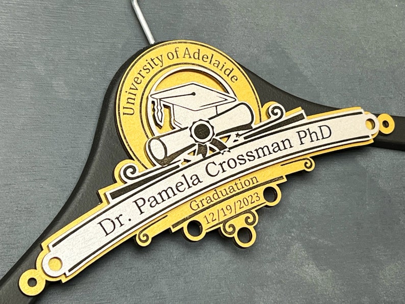 May include: Personalized PhD Hanger, PhD Gift, Personalized PhD Graduation Gift, Doctorate Graduation Gift, Hooding Ceremony Gift, Doctoral Candidate Black wooden hanger with a gold and silver inscription. The inscription reads "University of Adelaide, Dr. Pamela Crossman PhD, Graduation 12/19/2023". The inscription is decorated with a graduation cap, diploma, and scroll.